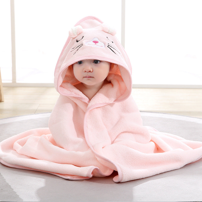 A baby is sitting on a light-colored mat, wrapped in a soft, pink hooded blanket with a cute animal face and whiskers design. The room has a bright and airy ambiance.