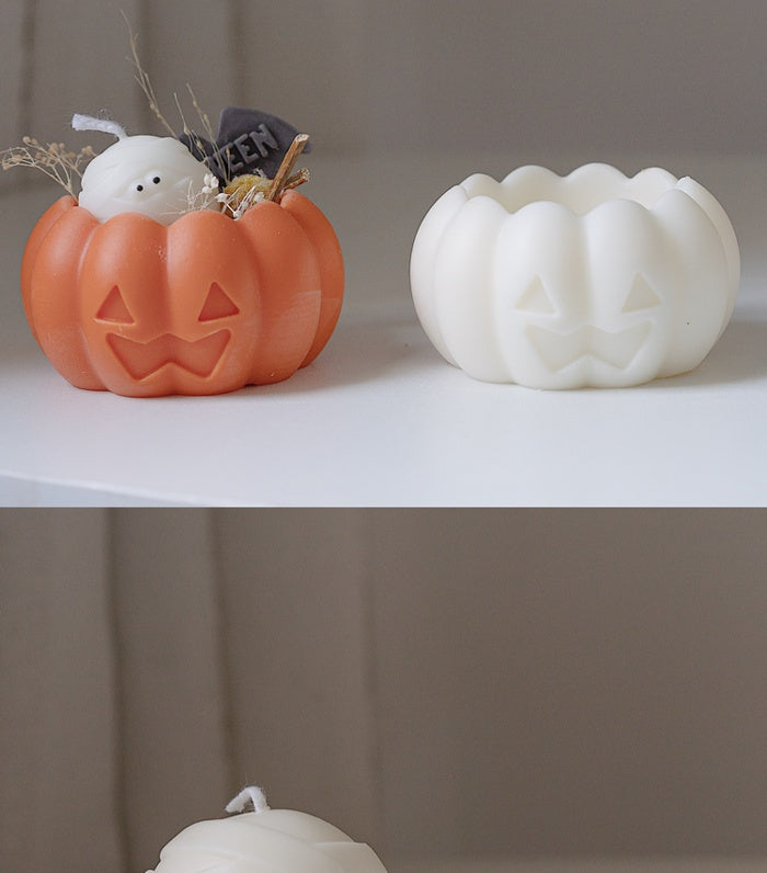 Two pumpkin-shaped candles: one orange with a Halloween decoration and ghost, the other white. Both have carved faces and sit on a light surface against a neutral background.