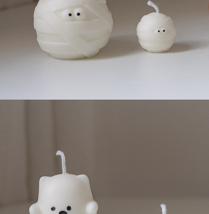 Two wax candles shaped like a mummy and a bear on a white surface. The top half shows mummy-shaped candles with eyes, while the bottom shows a bear candle with facial features.