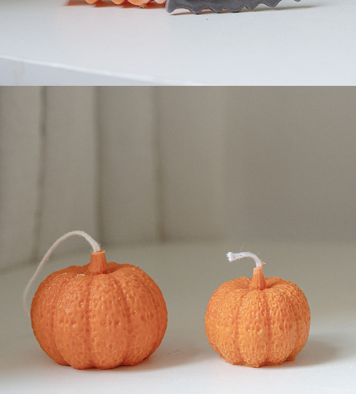 Two small orange pumpkin-shaped candles with textured surfaces sit on a white surface. Each candle has a white wick. The background is softly blurred, creating a cozy atmosphere.