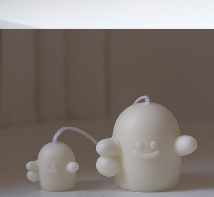 Two small white ghost-shaped candles with smiling faces and tiny arms. The larger one is slightly taller and both have wicks on top, set against a neutral background.