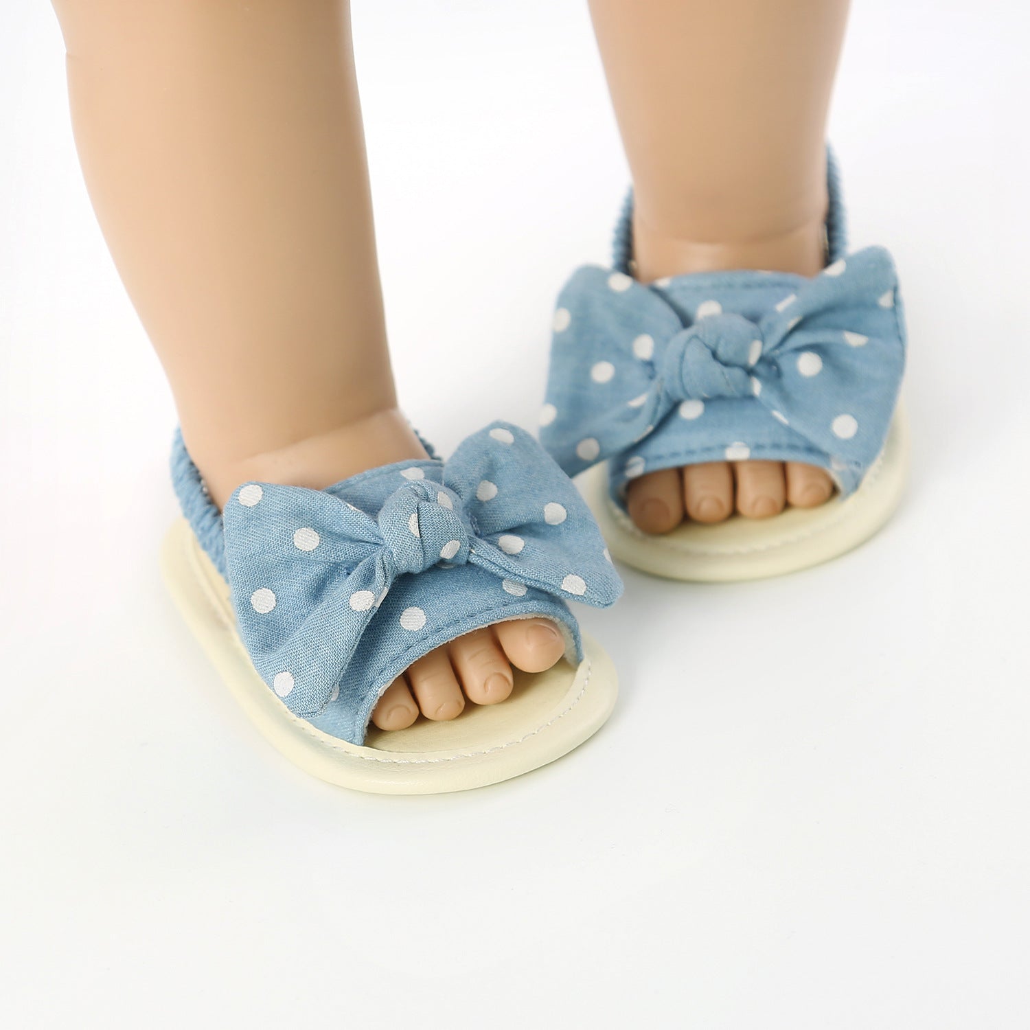 Baby feet wearing blue polka dot sandals with large bows. The sandals are open-toed and have stretchy fabric around the ankles.