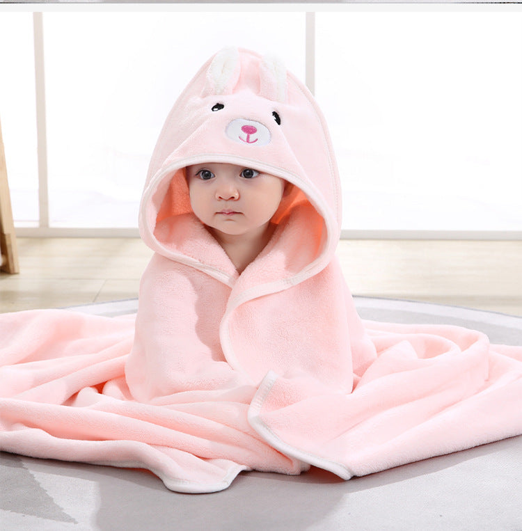 A baby wrapped in a soft pink hooded blanket with rabbit ears and face design, sitting on a rug in a bright room.