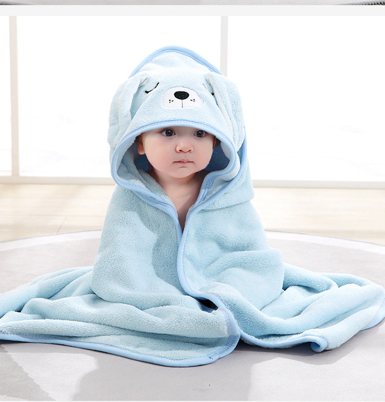 Baby wrapped in a cozy, light blue bear-themed hooded blanket, sitting on a soft, round rug in a well-lit room with large windows in the background.