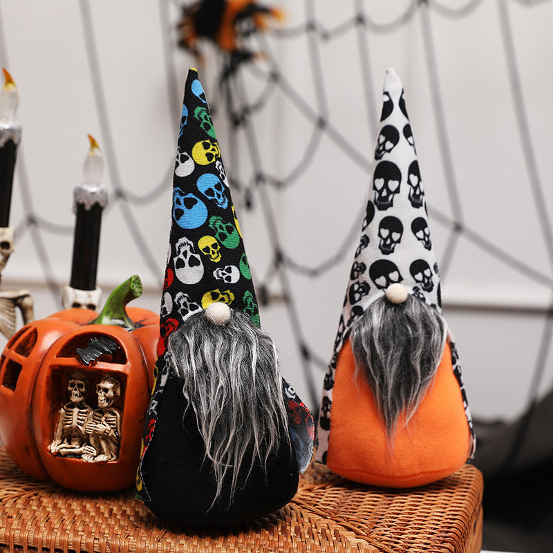 Two gnome figurines with long gray beards and pointed hats decorated with colorful and black skull patterns stand on a wicker table. They are surrounded by Halloween decor, including a pumpkin with skeletons and skeleton-themed candles. A spider web hangs in the background.