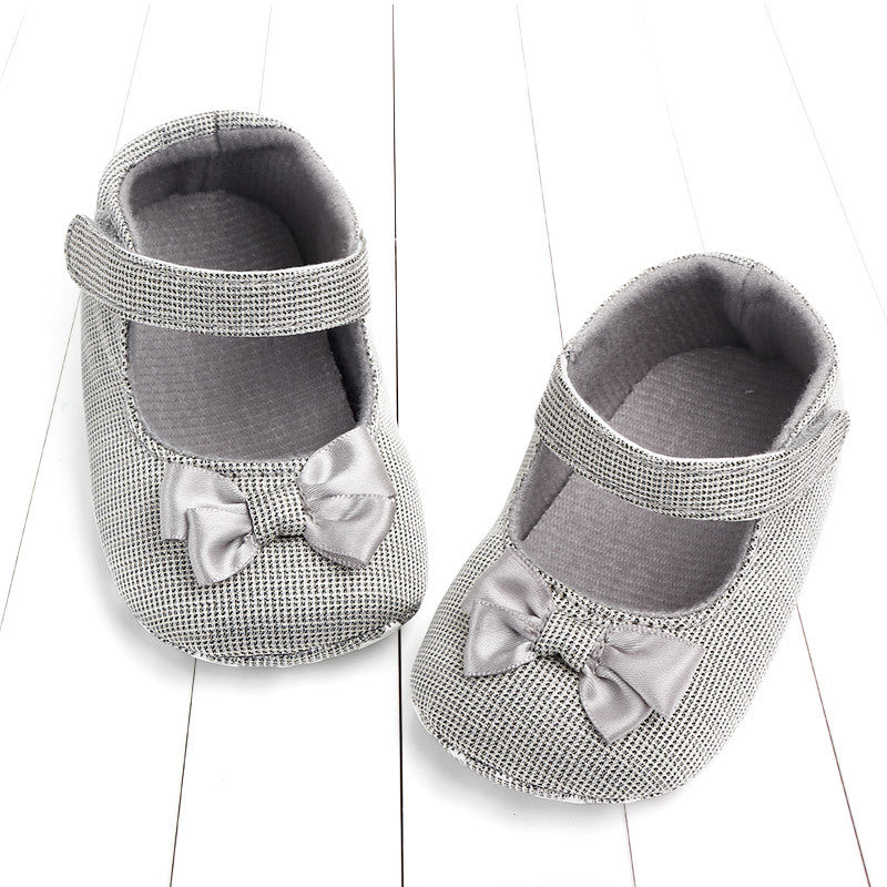Gray baby shoes with a textured pattern, featuring bow accents and Velcro straps, placed on a white wooden surface.