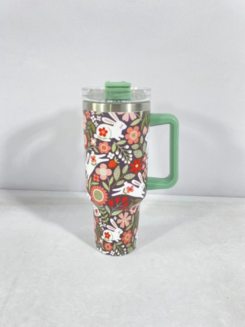 A floral tumbler with a green handle and lid, adorned with white rabbits and colorful flowers on a black background, standing on a light gray surface.