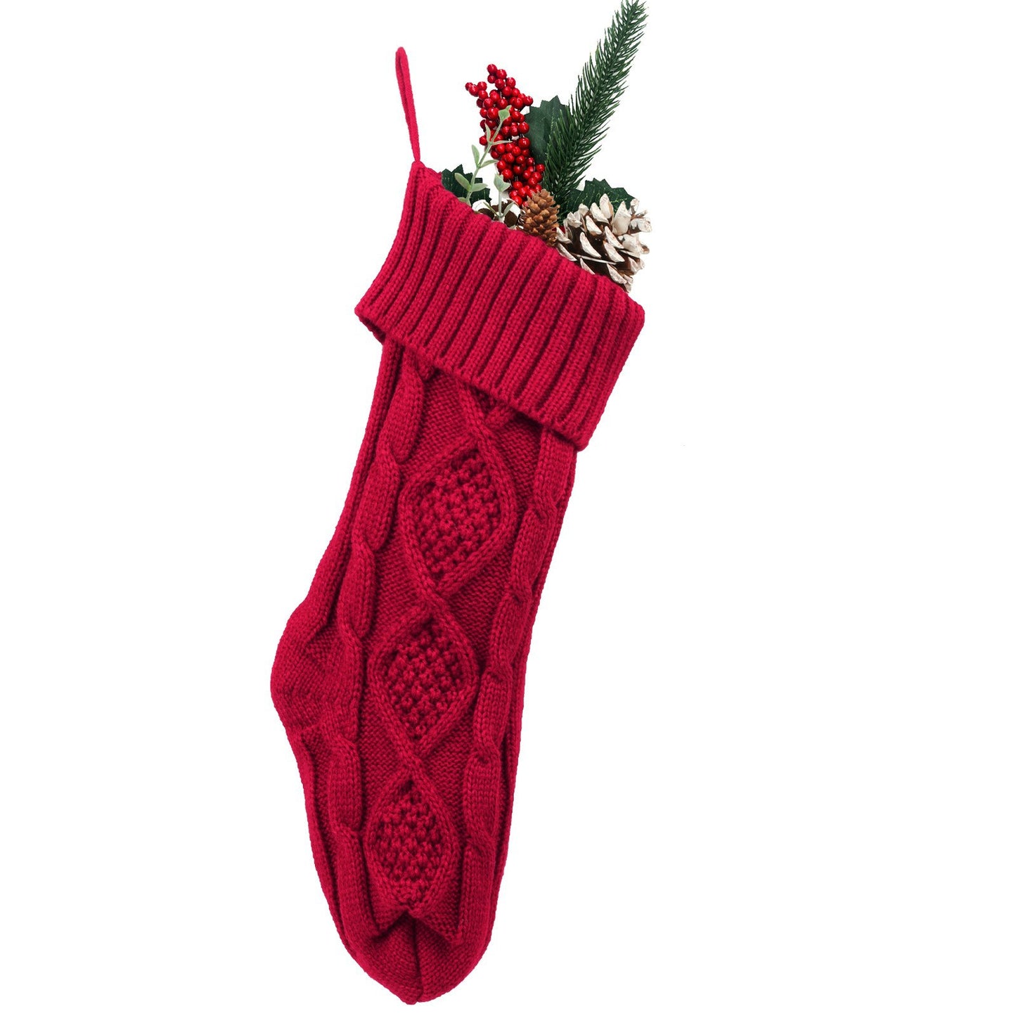 A red cable-knit Christmas stocking filled with red berries, pine branches, and pinecones, set against a plain white background.