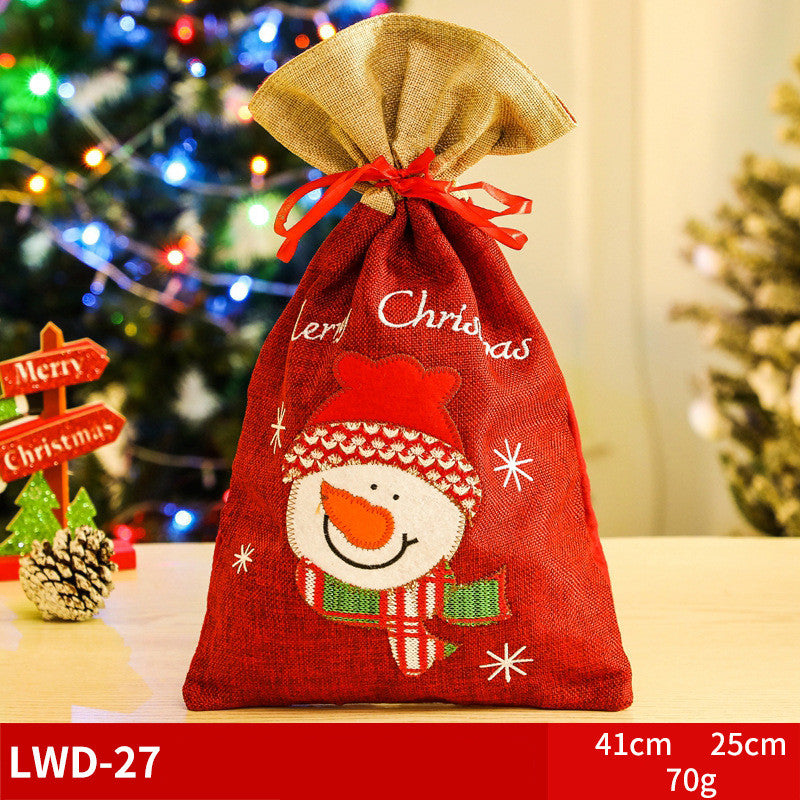 Festive red gift bag with a snowman design, burlap top, and red bow, set in front of a lit Christmas tree. Dimensions and weight listed as 41cm x 25cm x 70g.