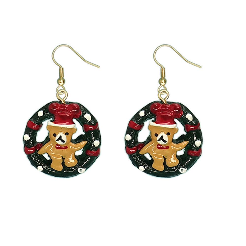 Festive earrings featuring a teddy bear with a red hat inside a green wreath adorned with red ribbons and white accents. The earrings have gold hooks.