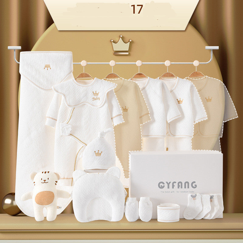 A collection of baby clothes and accessories, including onesies, hats, mittens, socks, a blanket, and a plush toy bear. All items feature white and beige tones with crown motifs.