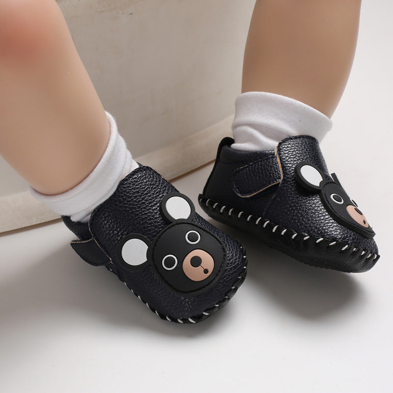 Baby wearing black leather shoes with cute bear designs on the front, paired with white socks. The shoes have a textured finish and white stitching along the sole.