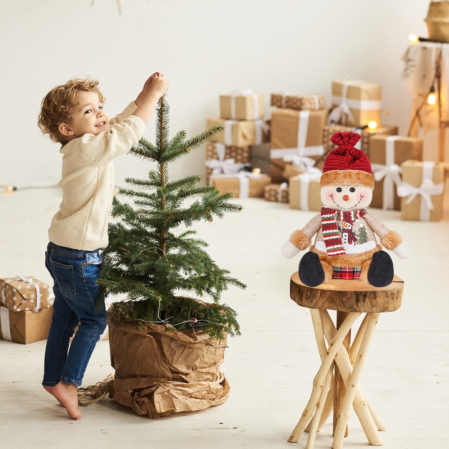 A young child decorates a small Christmas tree in a cozy setting with wrapped gifts and a plush snowman toy on a wooden stool nearby.