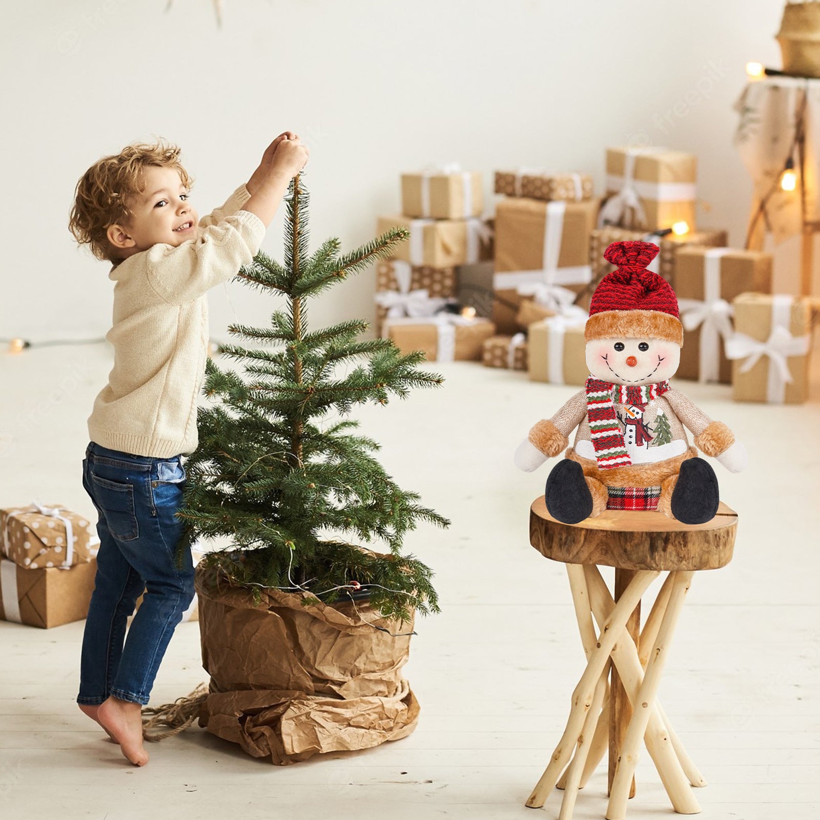 A young child decorates a small Christmas tree in a cozy setting with wrapped gifts and a plush snowman toy on a wooden stool nearby.