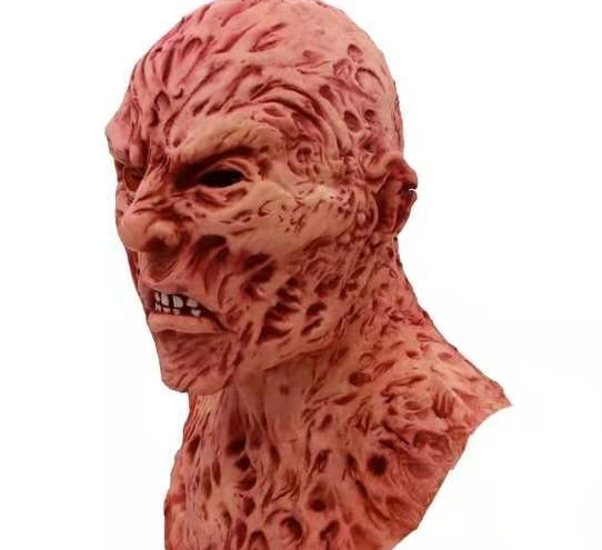 Rubber mask depicting a menacing, scarred face with bumpy, textured skin and a fierce expression. The reddish tone and exaggerated features create a monstrous appearance.