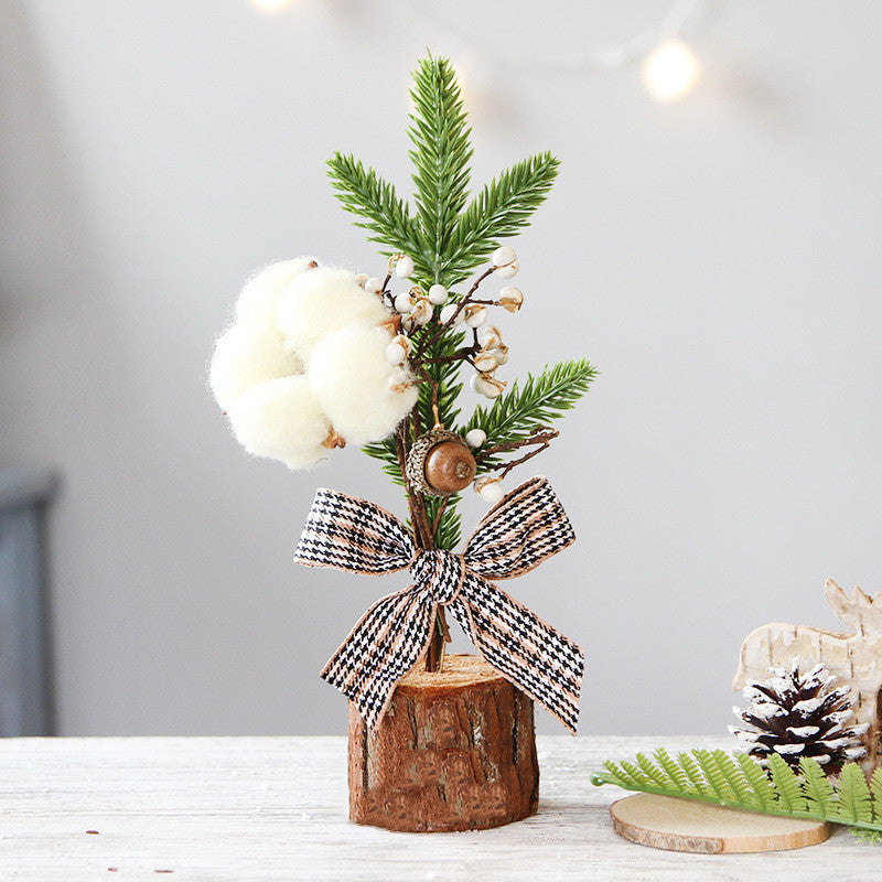 Miniature holiday decoration with pine branches, cotton tufts, and berries in a wooden base. Decorated with a black and white checkered bow, pinecone, and fern nearby.