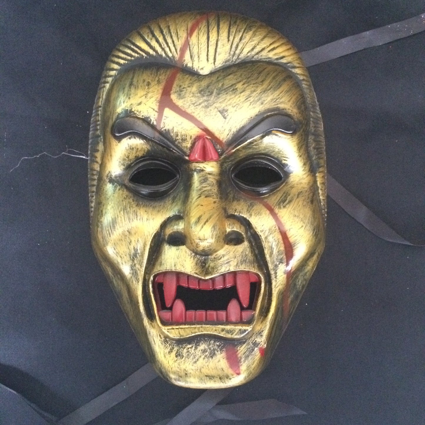 A golden, demonic mask with red scars, sharp fangs, and exaggerated features on a dark background, featuring prominent brow ridges and black eye holes.