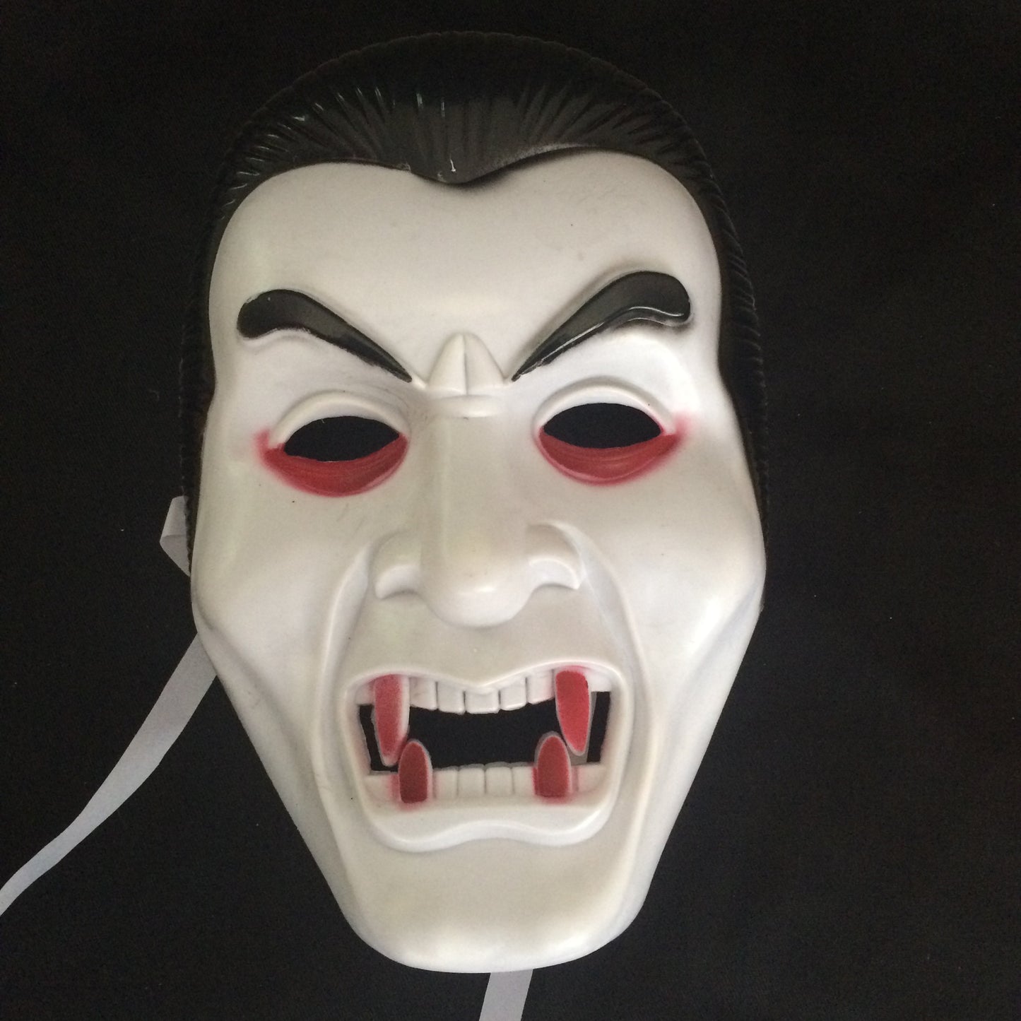 A vampire mask with exaggerated fangs, arched eyebrows, and red accents around the eyes, set against a dark background.