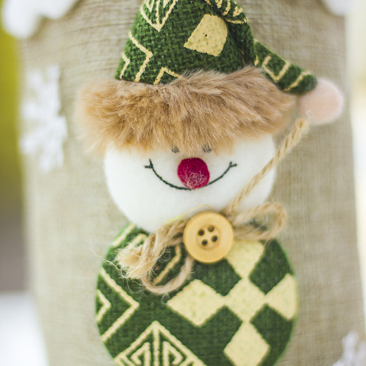 Festive snowman decoration with a fluffy hat, red nose, and smile. It wears a green patterned sweater and button, with a rope scarf, against a beige background.