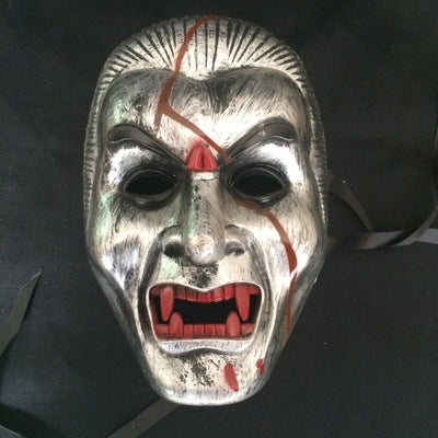 A silver vampire mask with black eye holes, red fangs, and blood-like red streaks across the face, set against a dark background.
