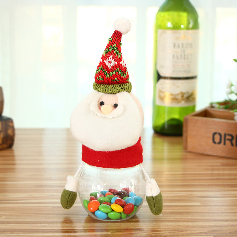Festive Santa Claus candy jar with a knitted hat, holding colorful candies inside a transparent base, placed on a wooden table. Background includes decor items and a wine bottle.