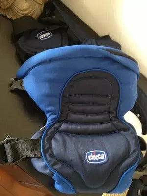 A blue and black Chicco baby carrier is placed on a dark surface, featuring padded straps and a secure buckle, with its brand logo prominently displayed at the center.