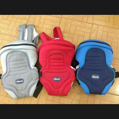 Three baby carriers are displayed on a wooden floor. They are in gray, red, and blue, each with adjustable straps and a logo in the center front.