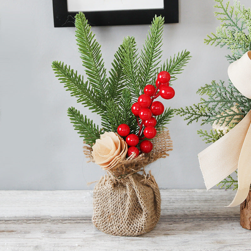 Small burlap-wrapped arrangement with pine branches, red berries, and a beige flower on a wooden surface, set against a neutral background.
