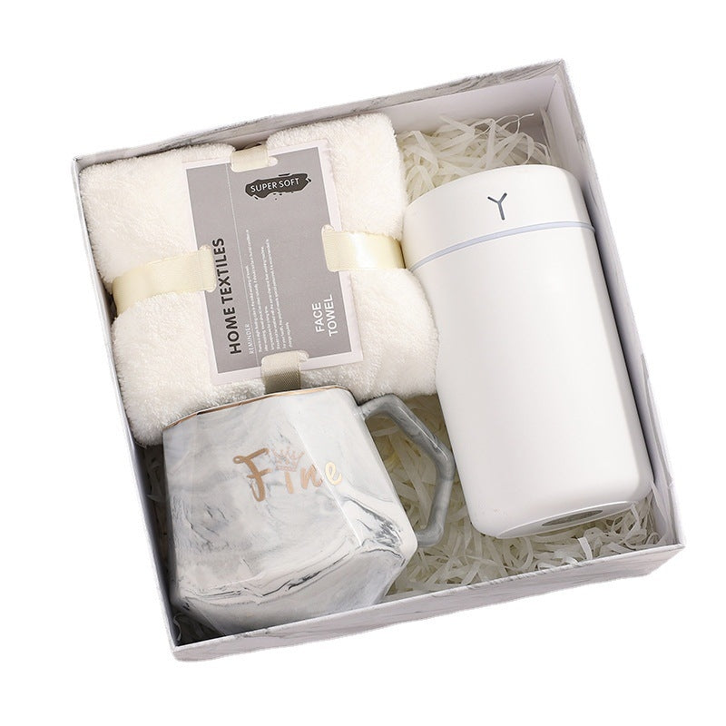 Gift box containing a folded white face towel, a white insulated tumbler, and a marble-patterned mug with the word "Fine" in gold, set on crinkled paper.
