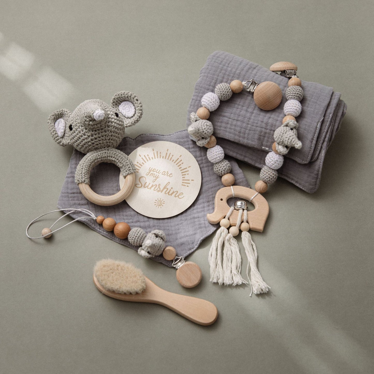 A crochet elephant rattle, wooden teething clip, soft gray cloth, baby brush, and a wooden plaque with "you are my sunshine" are delicately arranged on a light surface.