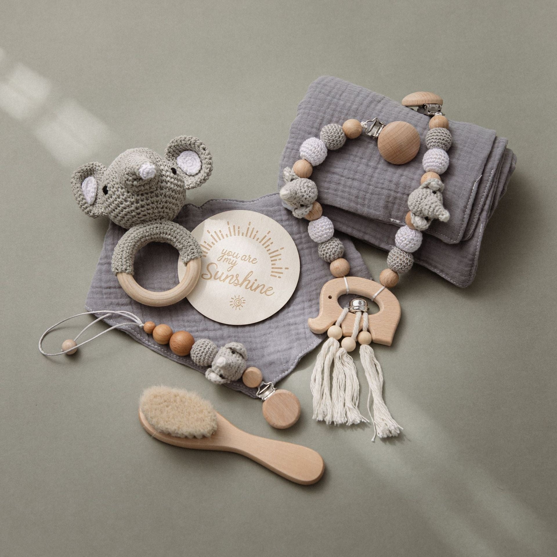 A crochet elephant rattle, wooden teething clip, soft gray cloth, baby brush, and a wooden plaque with "you are my sunshine" are delicately arranged on a light surface.