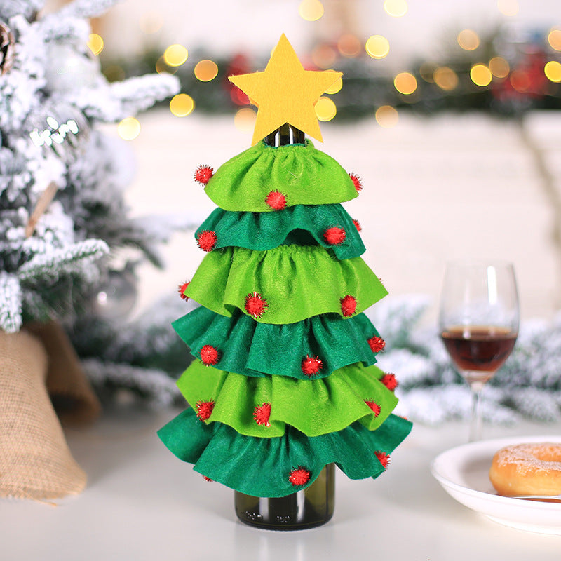 Festive bottle cover resembling a Christmas tree with layered green fabric, red pompoms, and a yellow star on top. Nearby are a wine glass and a plate with a pastry.