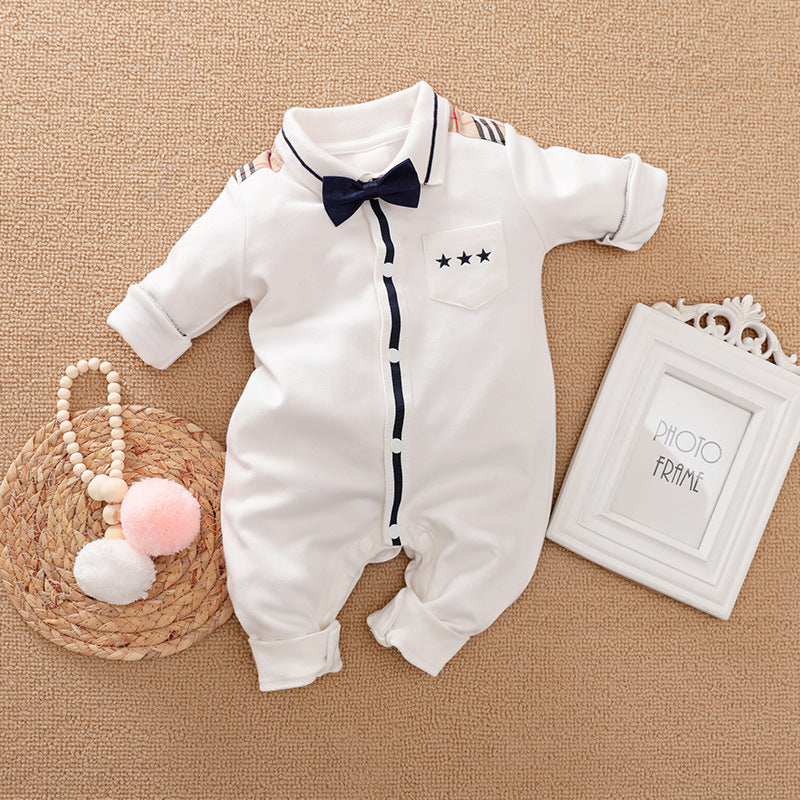 White baby onesie with navy trim, bow tie, striped shoulders, and star-embellished pocket lies on a beige surface. Nearby are a pink-and-white pom-pom accessory and an empty white photo frame.