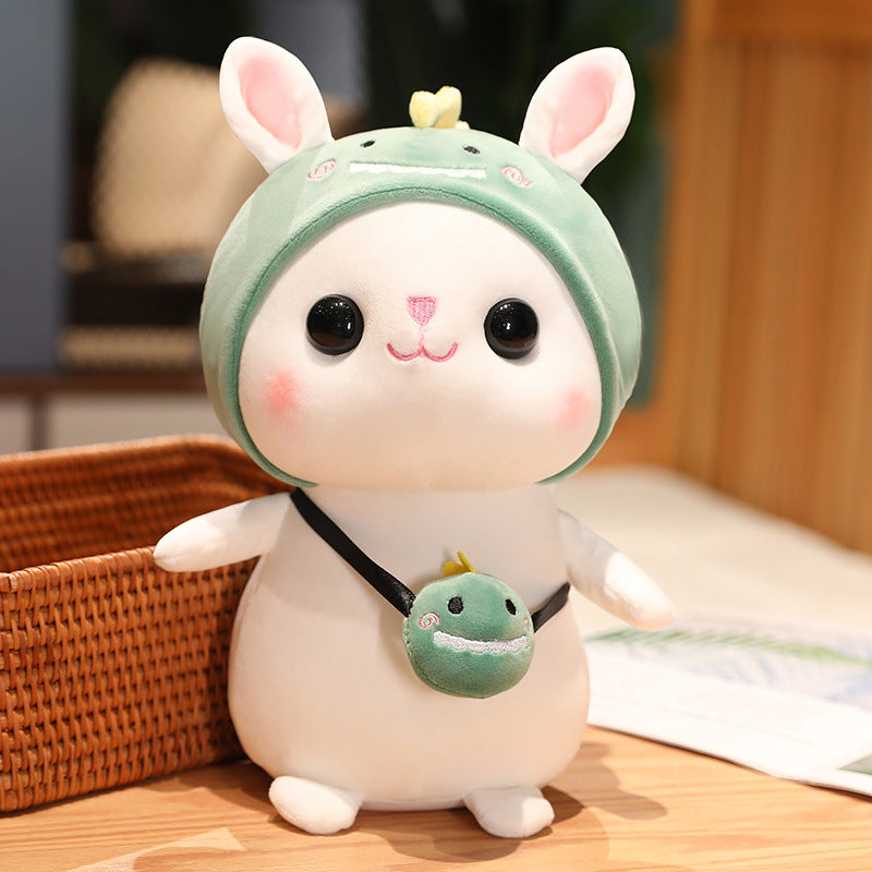 Cute plush toy rabbit with a green dinosaur hoodie and matching small bag. It has large eyes, pink cheeks, and is set on a wooden surface next to a wicker basket.
