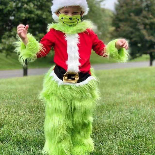 Child dressed as the Grinch in a Santa hat and red jacket with green furry pants, standing on grass, mimicking the character's playful pose in an outdoor setting.