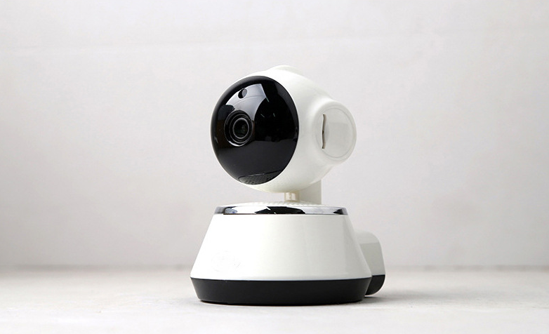 A small, modern white and black security camera with a rounded design is positioned on a light surface, angled slightly to the left. The camera lens is prominently visible.