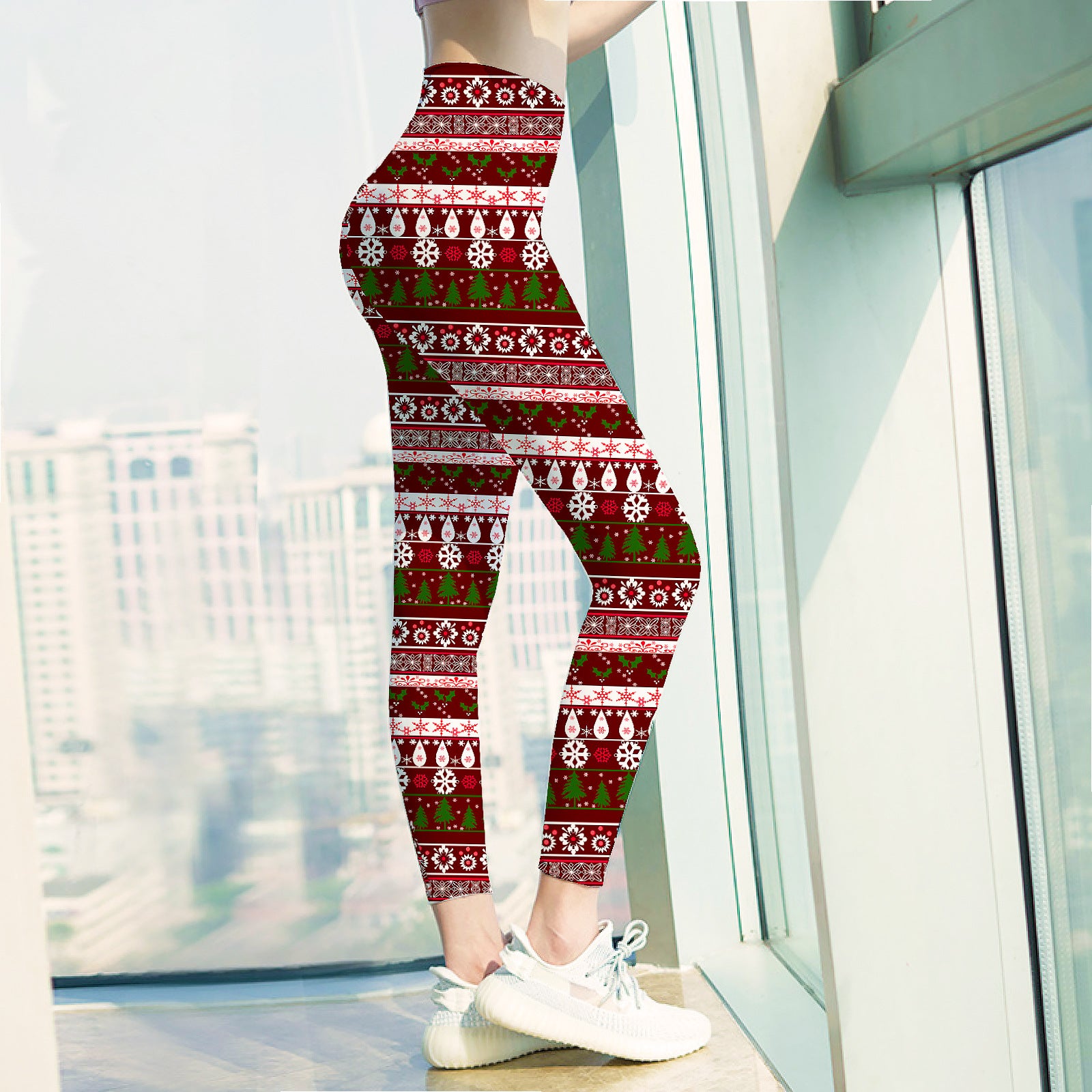 Person wearing festive red leggings with snowflakes and trees, standing by large windows with a city view. Paired with white sneakers.