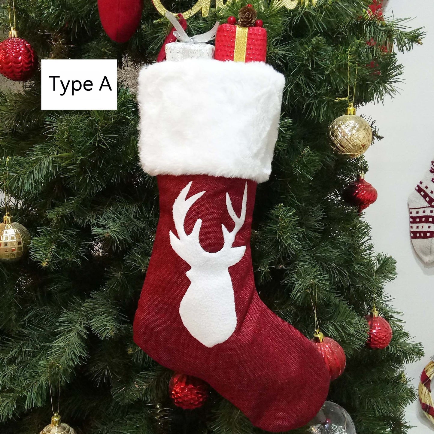 Red Christmas stocking with a white faux fur cuff and reindeer silhouette hangs on a decorated tree. It's filled with wrapped gifts. Label reads "Type A."