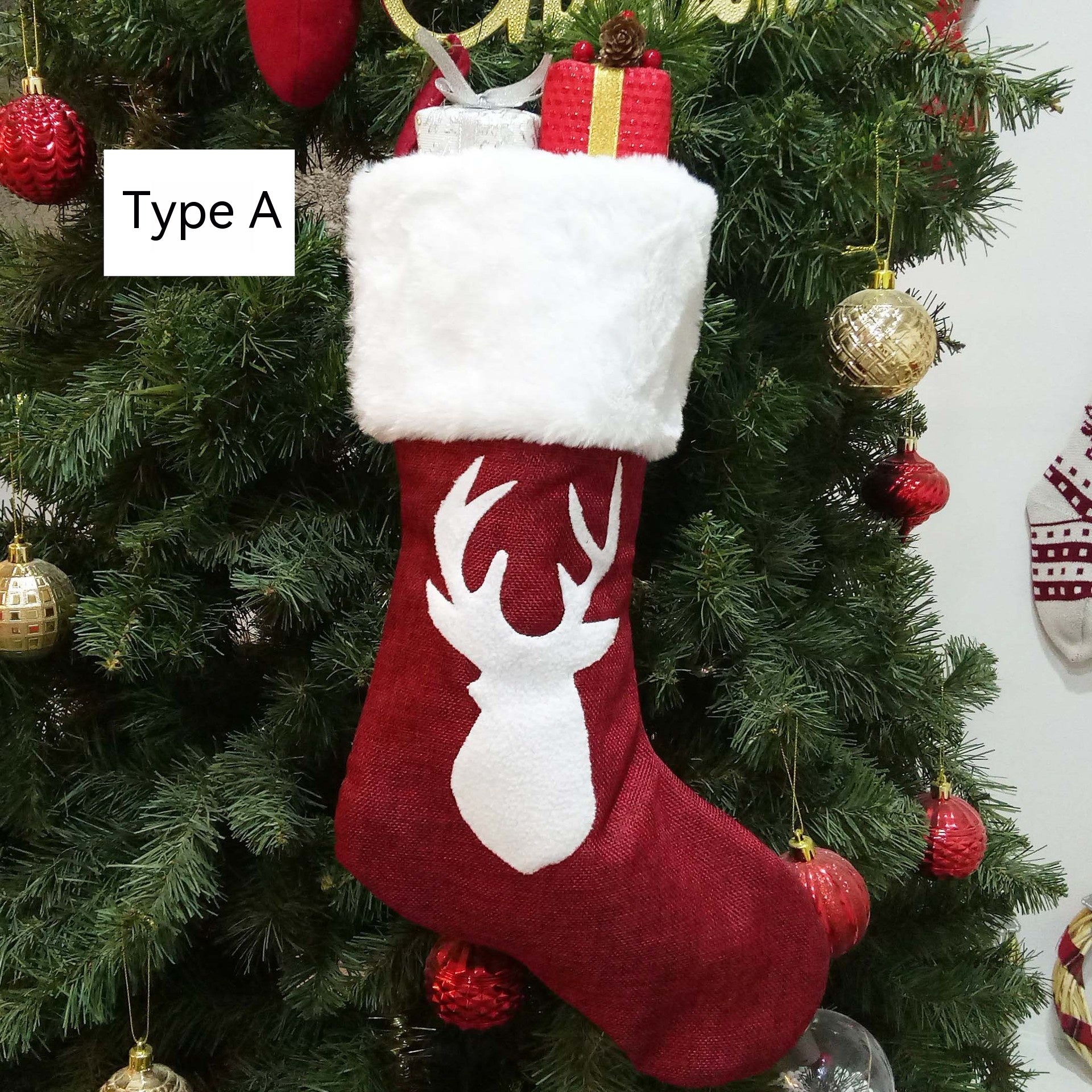 Red Christmas stocking with a white faux fur cuff and reindeer silhouette hangs on a decorated tree. It's filled with wrapped gifts. Label reads "Type A."