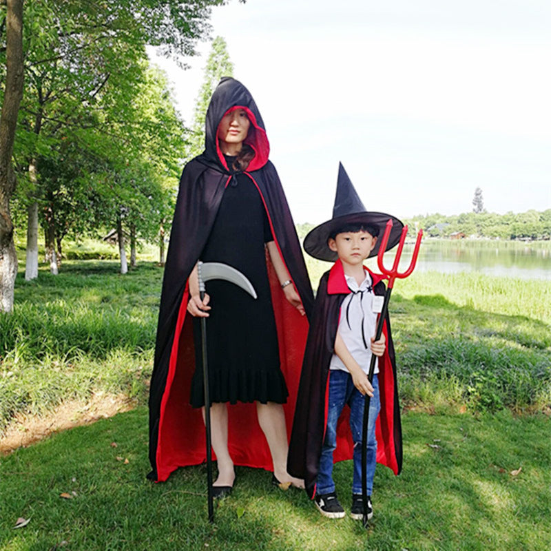 A woman and child stand outdoors dressed in Halloween costumes. The woman wears a black cloak with red lining and a hood, holding a scythe. The child wears a black witch's hat and cloak, holding a red trident. They are in a lush park with trees and a lake visible in the background.