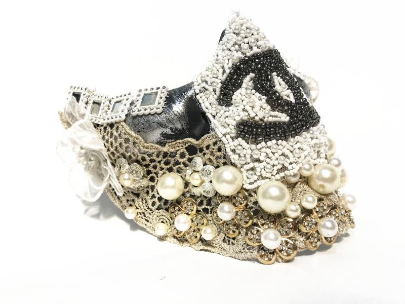 Ornate shoe adorned with intricate lace, pearls, rhinestones, and elaborate bead patterns, including an abstract motif on a white background. The heel is covered with gems.