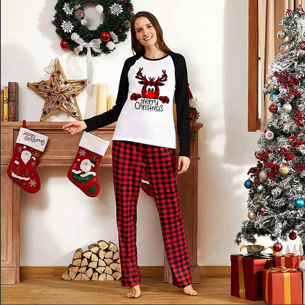 Smiling person in red plaid pajamas and a reindeer shirt stands by a decorated Christmas tree, stockings, and a wreath, surrounded by festive decorations and gifts.