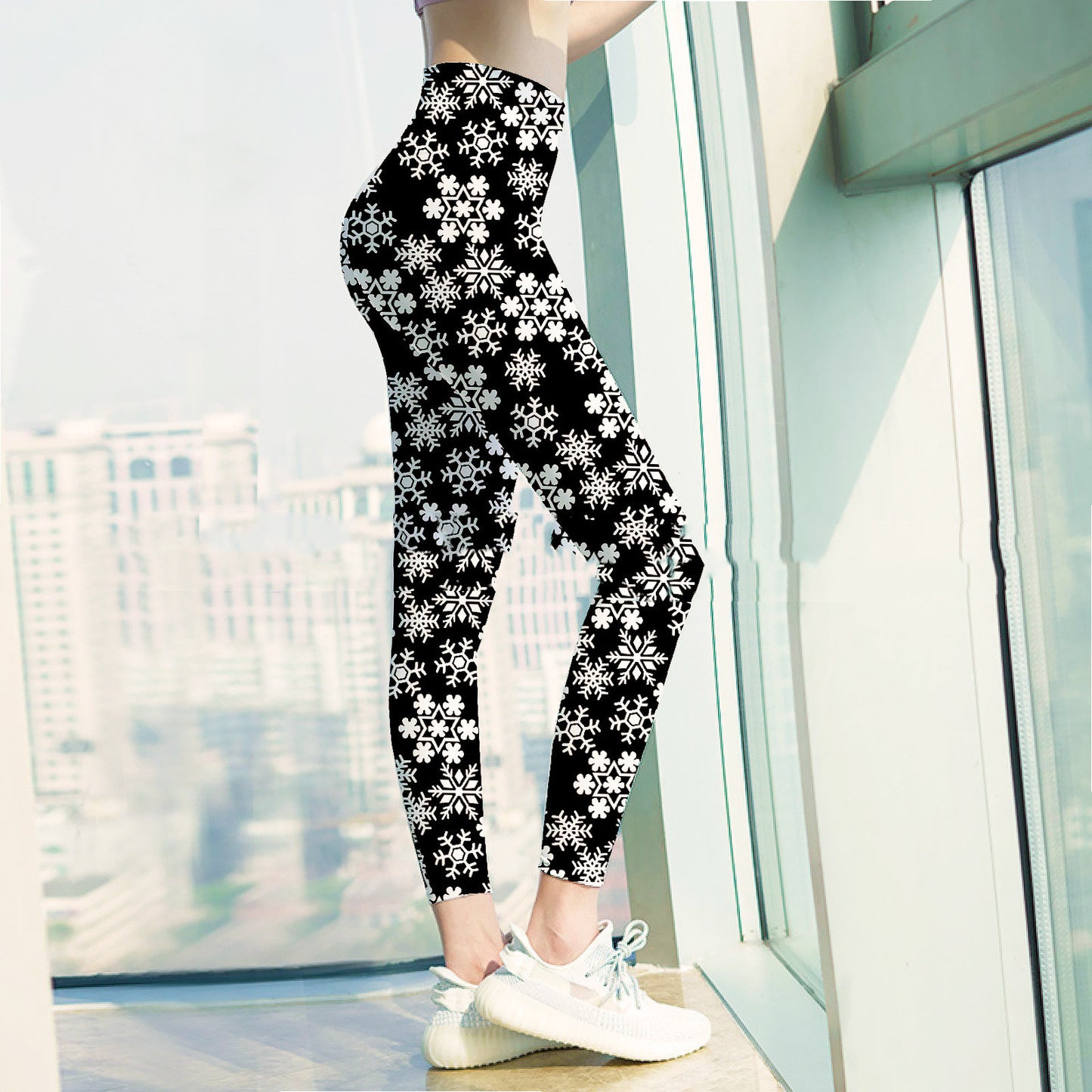 Person in high-waisted black leggings with white snowflake patterns, wearing white sneakers, standing by a large window with a cityscape view in the background.