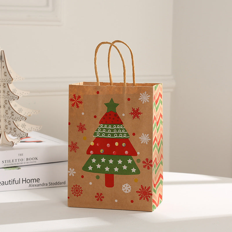 Brown gift bag with a colorful Christmas tree design and snowflakes, featuring twisted paper handles. Decorative tree shapes on a nearby book add to the festive setting.