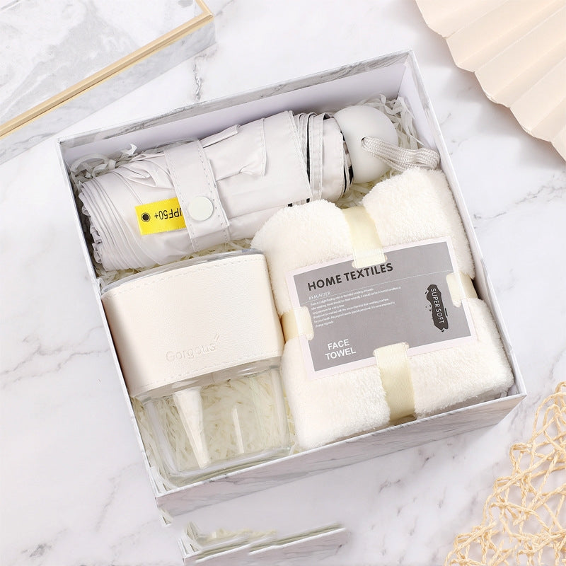 Gift set on marble surface featuring a compact white umbrella, a plush white face towel labeled "Home Textiles," and a clear glass jar with a white lid.