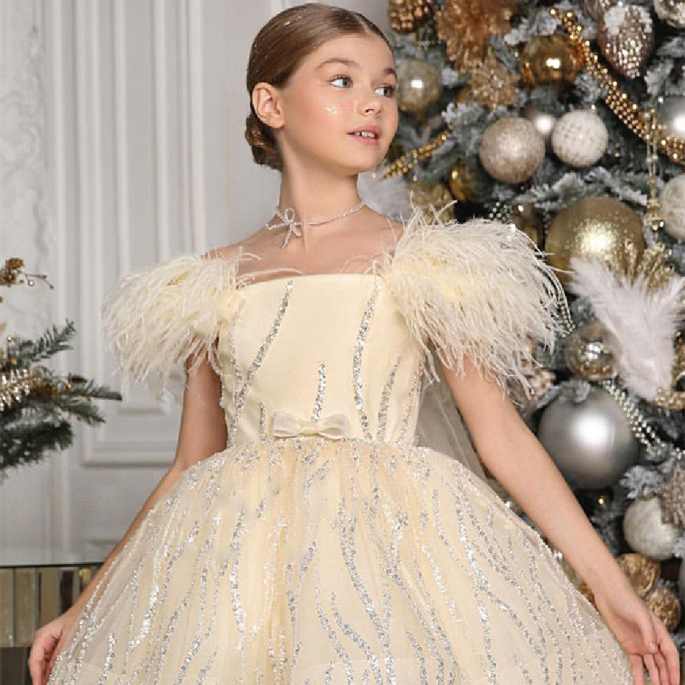 Girl in an elegant cream dress with feathered shoulders and silver accents, standing near a Christmas tree adorned with gold and silver ornaments in a decorated room.