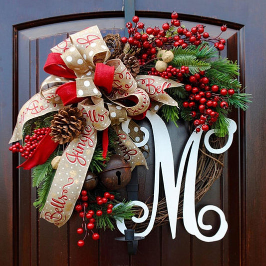 Festive wreath on a door with red berries, pinecones, green foliage, and a large ribbon featuring "Jingle Bells." A white letter "M" is prominently displayed in the center.