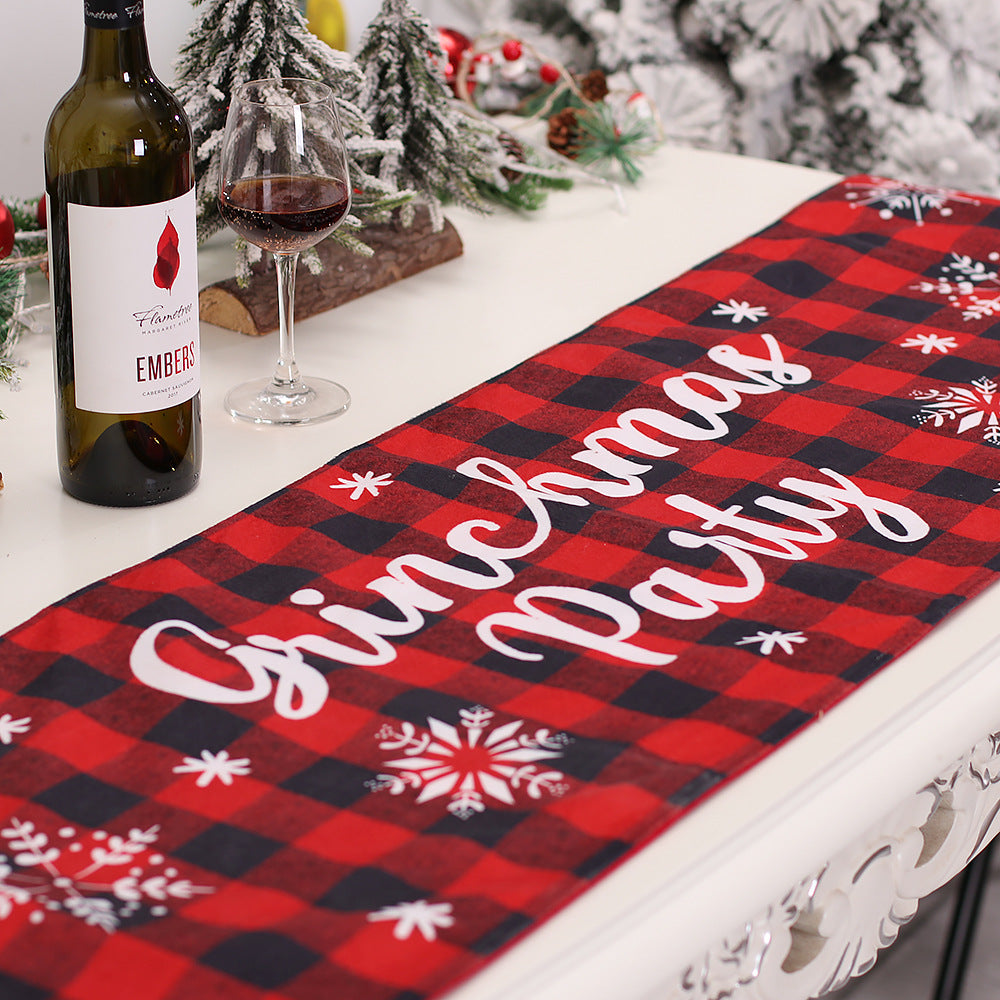 A festive table setup with a "Grinchmas Party" red and black plaid runner, a bottle and glass of wine, and snowy decor in the background, including pine branches.