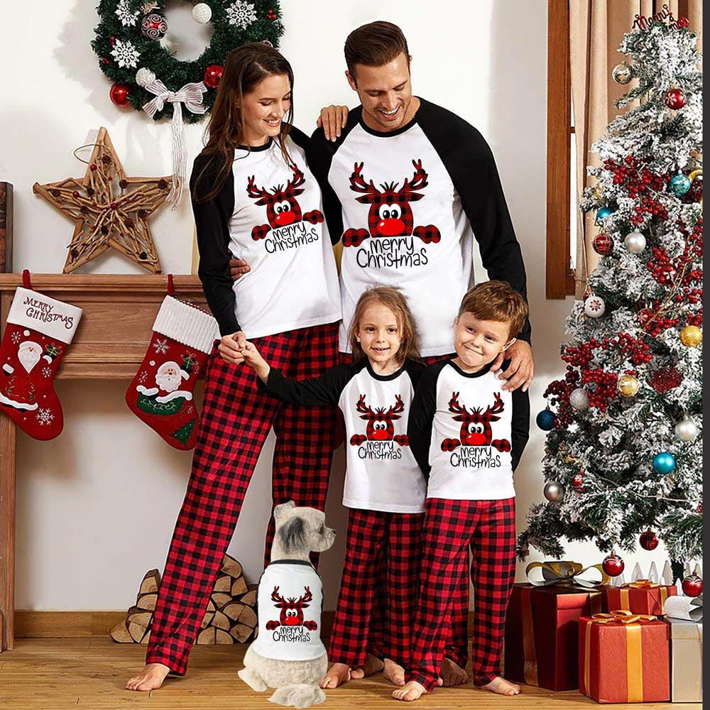 Family wearing matching "Merry Christmas" pajamas, standing by a decorated tree and stockings. A dog in matching attire sits by their feet. Festive room decor in the background.
