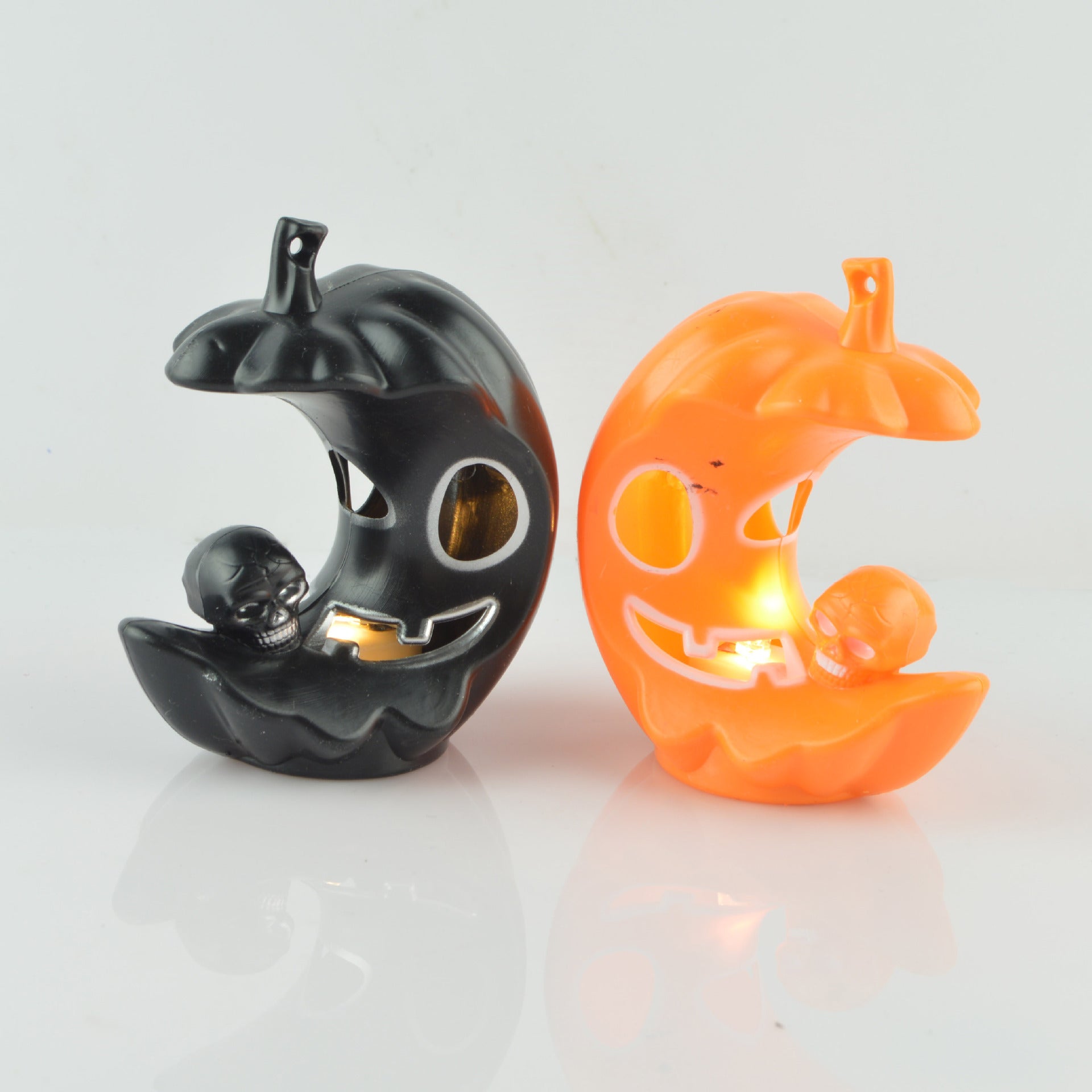 Two crescent-shaped pumpkin candle holders, one black and one orange, each with a grinning face and a small skull. Both contain lit candles, casting a warm glow.